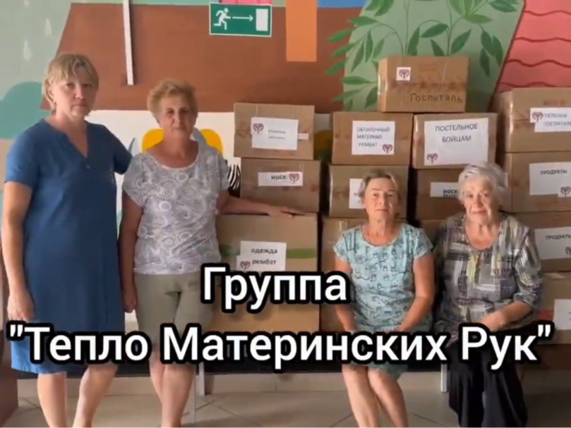 На изображении четыре женщины, стоящие и сидящие рядом с коробками. На переднем плане надпись «Группа „Тепло Материнских Рук“»..